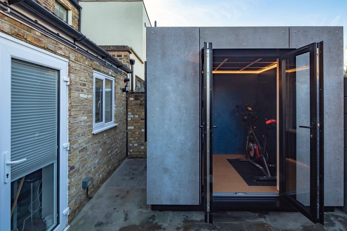 Concrete-look office and gym pod with both doors open, an exercise bike inside, beside the house