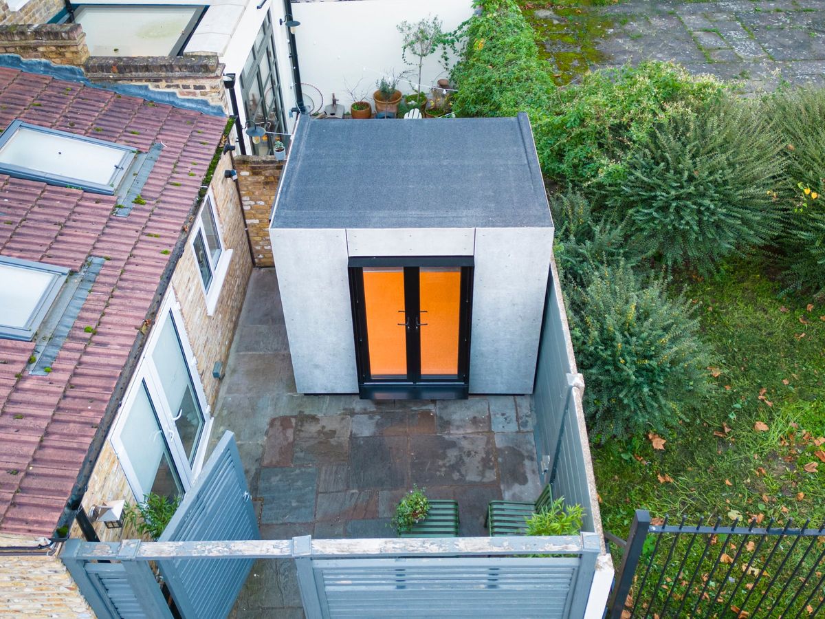 The concrete office pod seen from above in a small paved back yard, its orange-lit doors facing the house