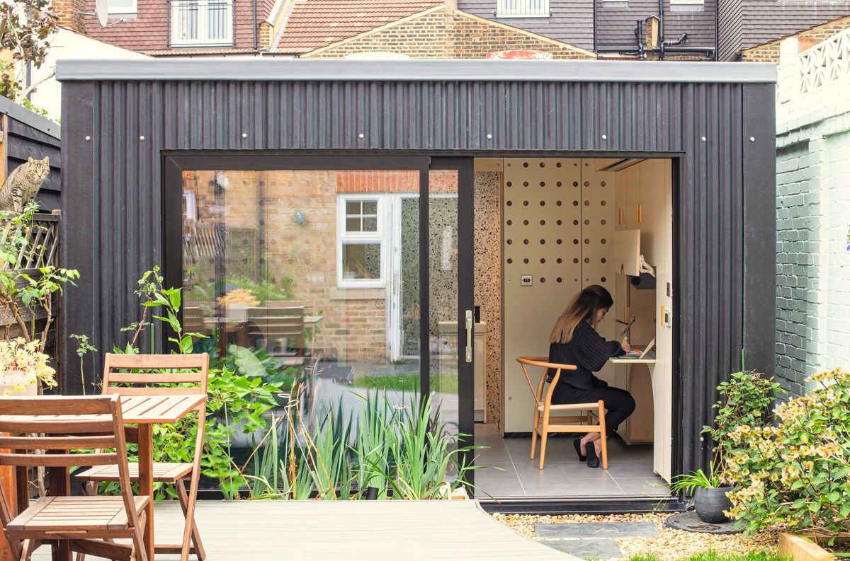 Dark-clad garden room annex in Walthamstow, its sliding door open and a woman working at the desk