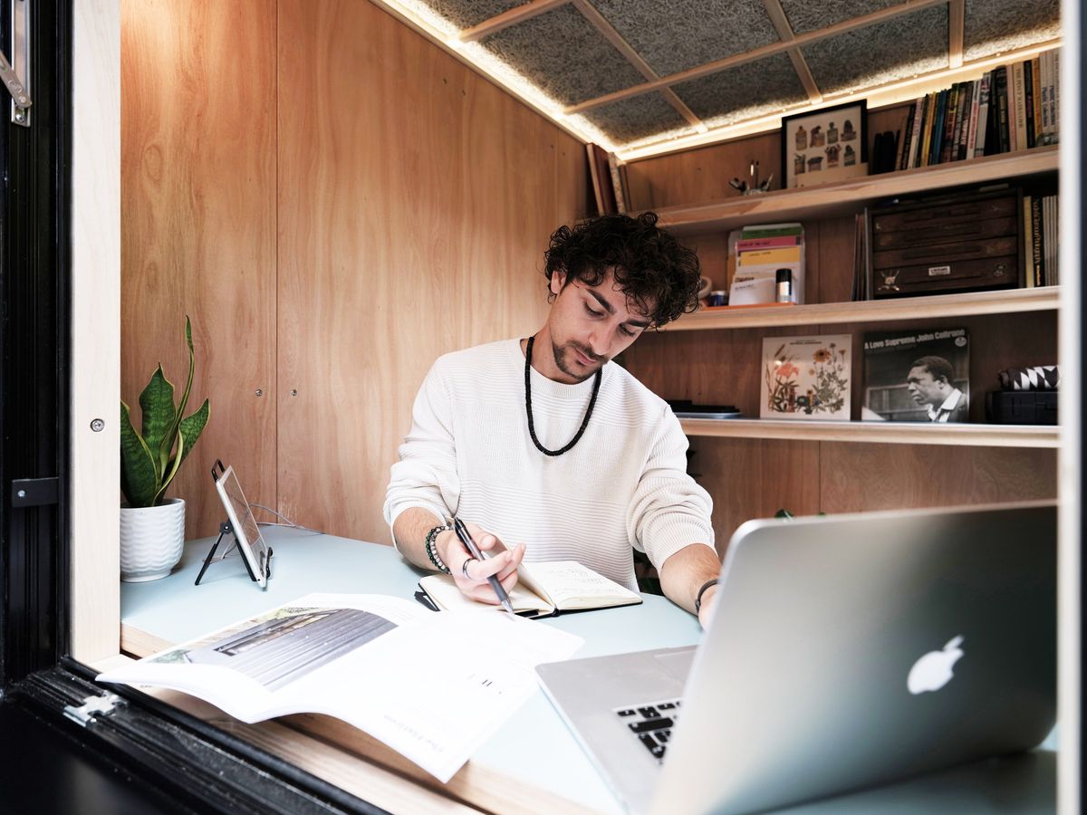Man working at the desk in the garden office pod, laptop and notebook