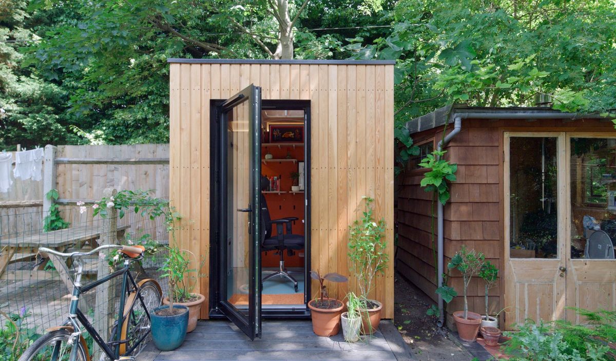 Tall, narrow larch-clad garden room office pod in a small Walthamstow garden, door open, next to an old shed