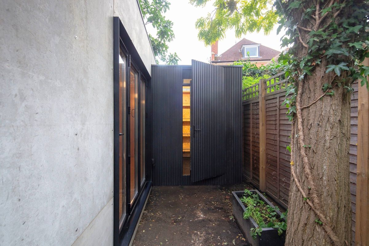 A path down the side of a garden studio to its clad storage room, the door open on the shelves