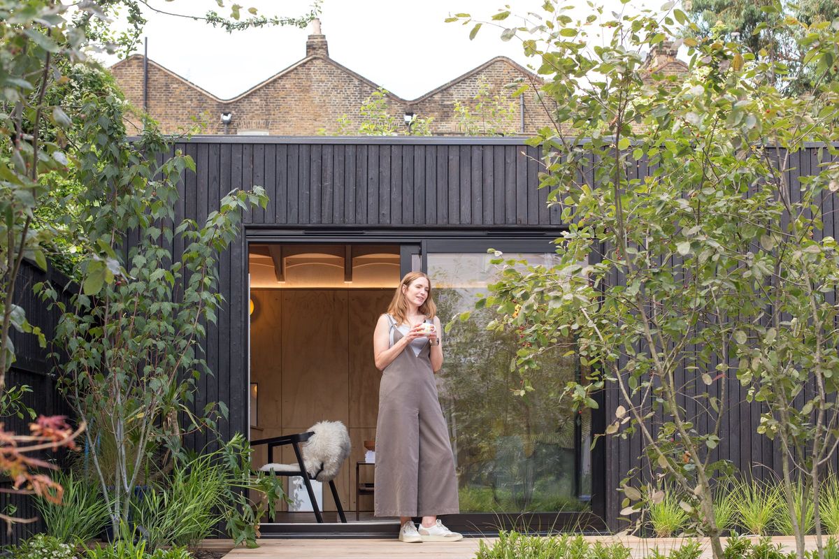 L-shaped garden studio clad in charred timber, its sliding door open, a woman standing in the doorway